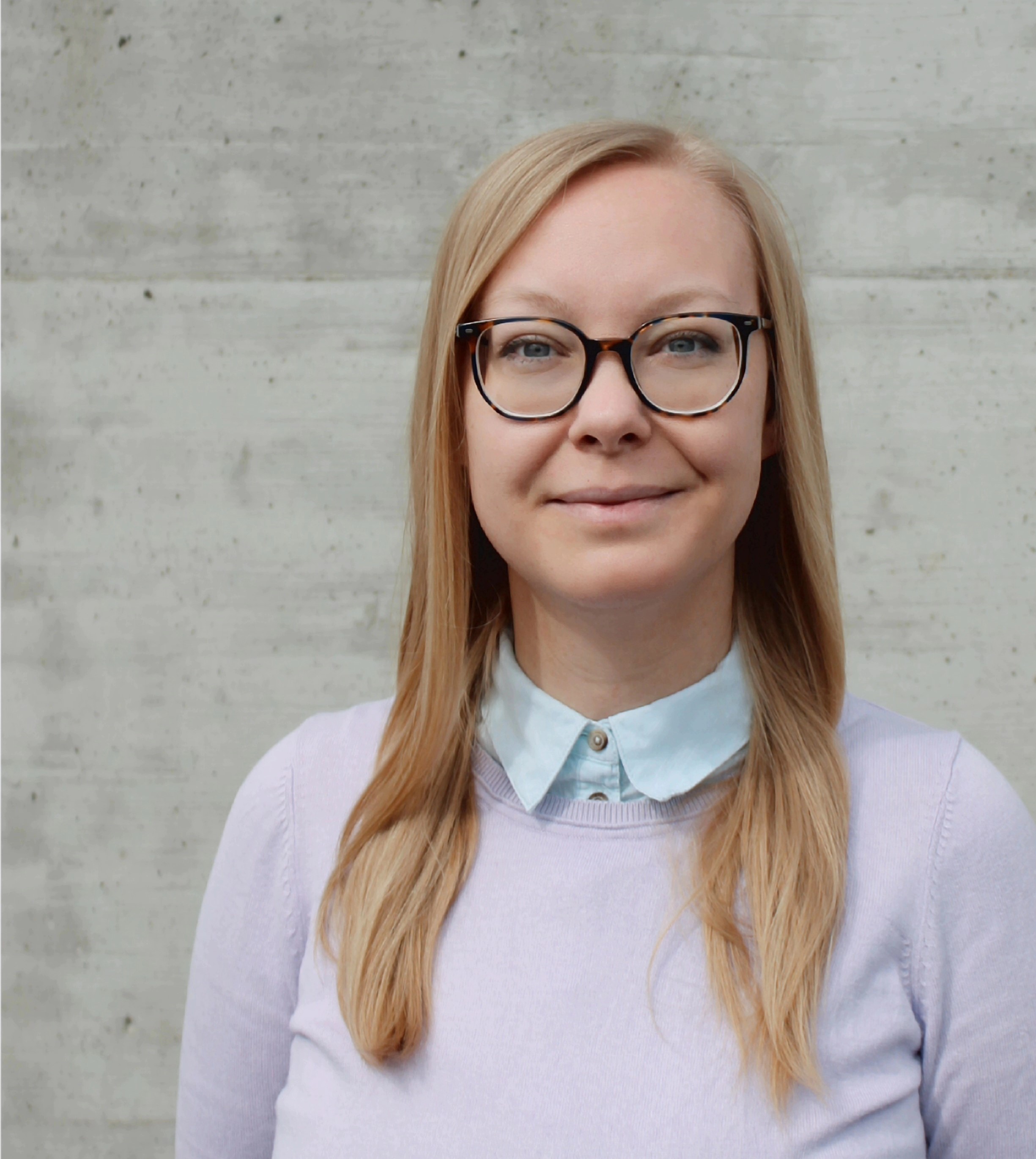 Michaela Joch ist eine weiße Frau Mitte 30, mit blonden 
langen Haaren und blauen Augen. Sie trägt eine Brille, eine hellblaue Bluse und darüber einen lila Pullover. Das
Foto ist vor einer Betonwand aufgenommen.